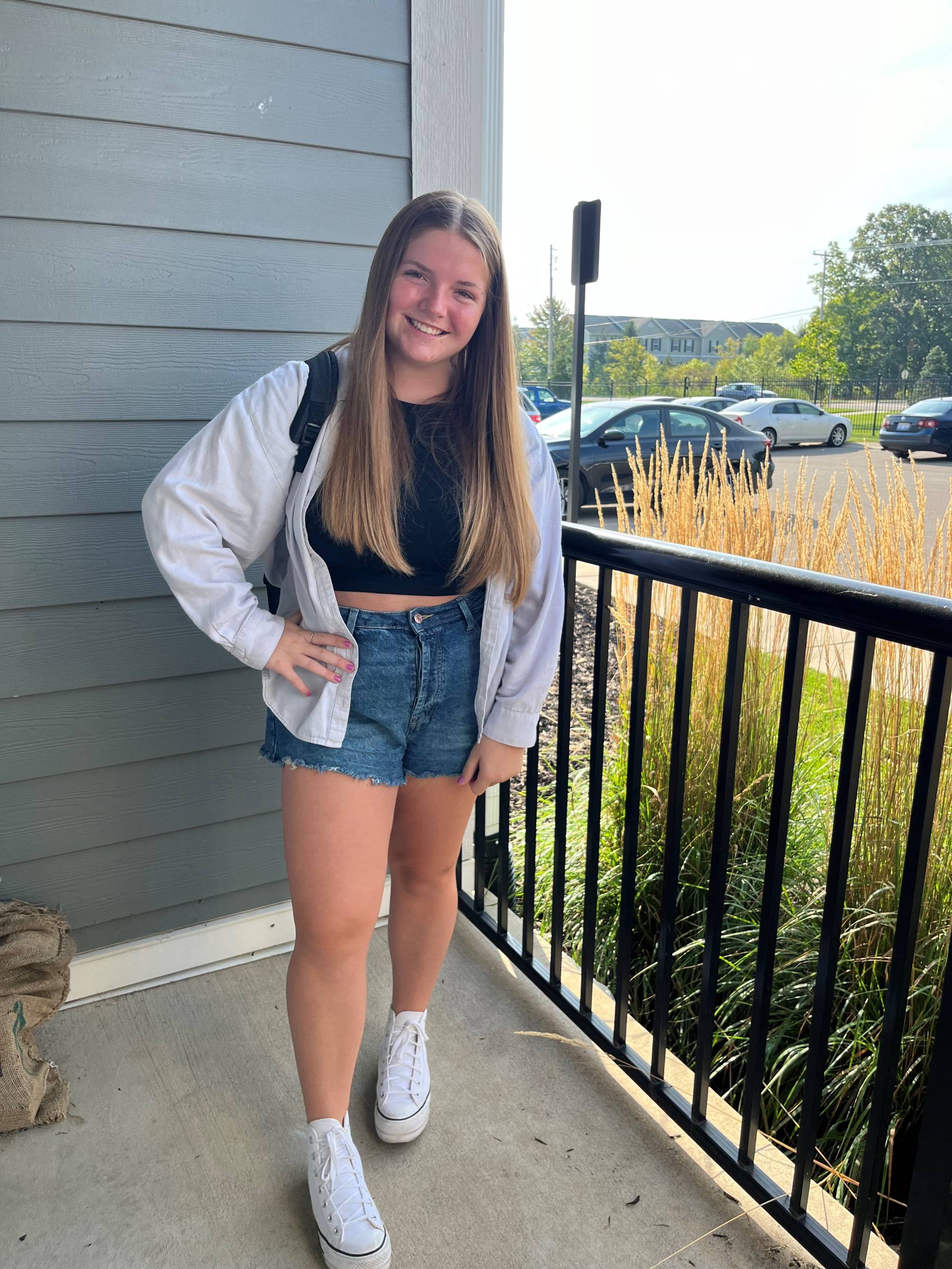 Girl with long blonde hair smiling at the camera. She is wearing jean shorts, a black top and a white jacket.
